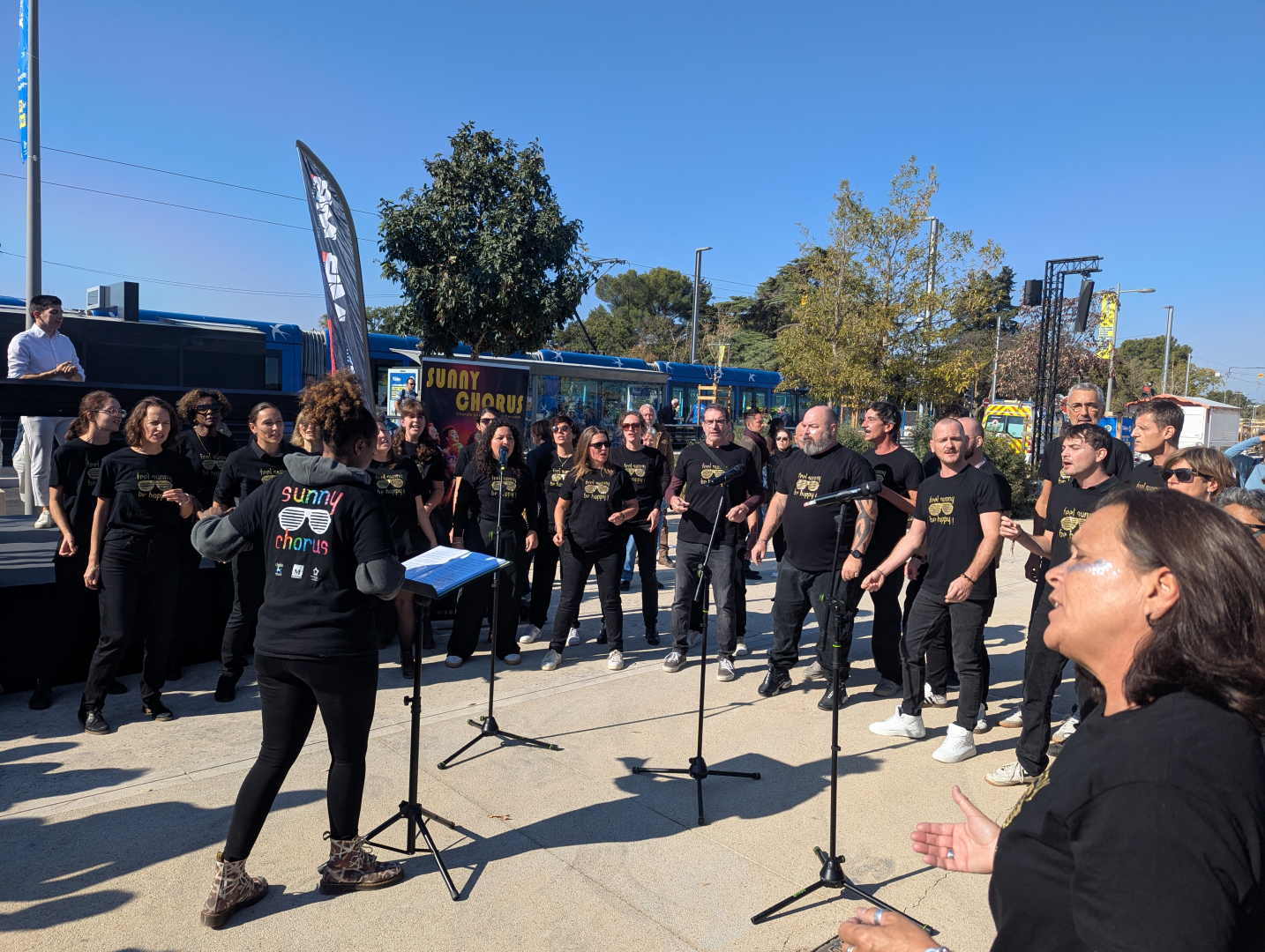 chorale Sunny Chorus en action devant rame de tramway micros sur pied ciel bleu arbre